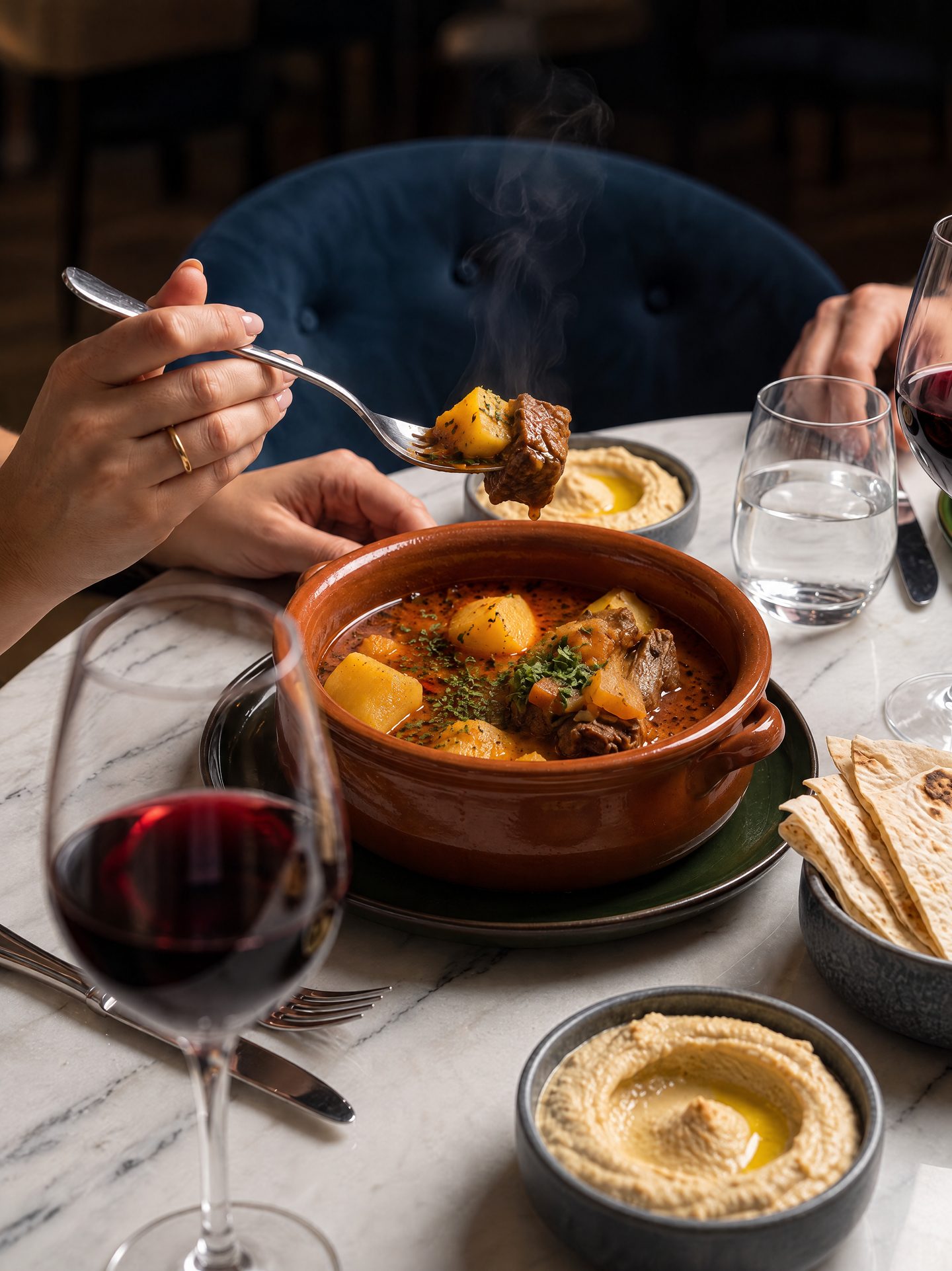 A steaming clay bowl of lamb and potato stew on a marble table, with dips, flatbread and red wine