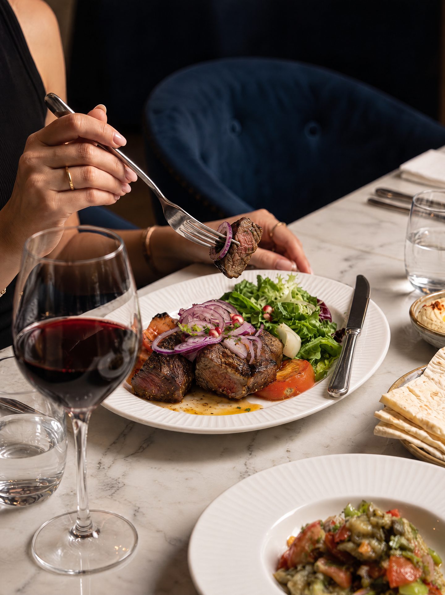 Grilled meat with salad and red wine on a marble table, with a guest reaching in with a fork