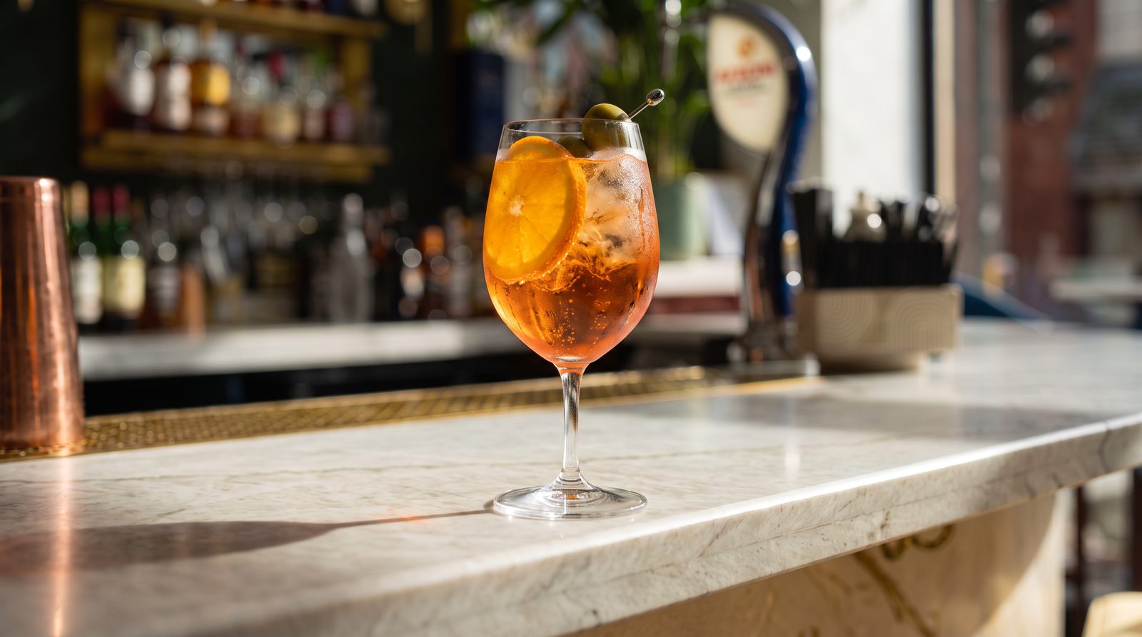 An Aperol Spritz with orange and an olive, in a wine glass on the marble bar
