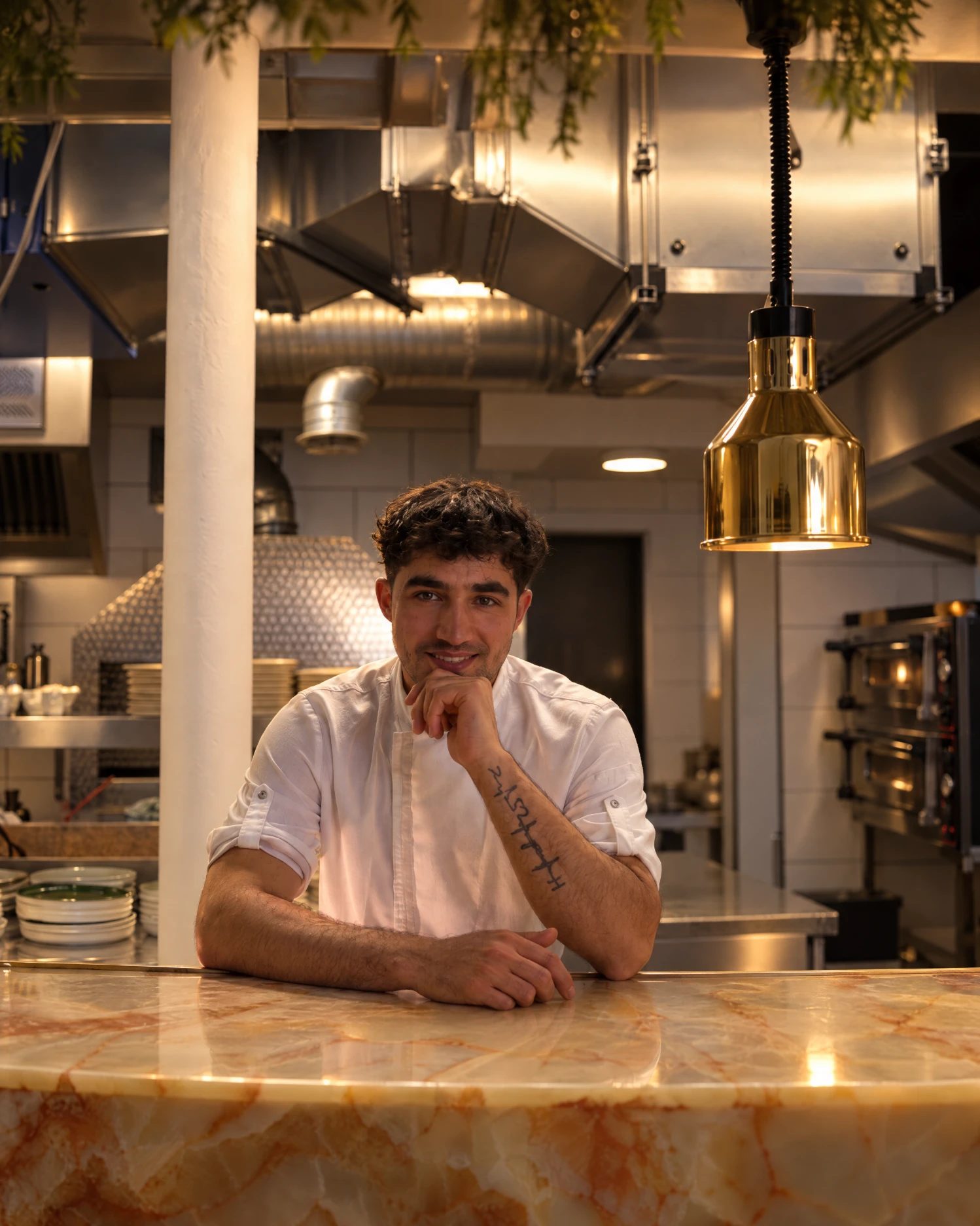 Amir in a white chef's shirt, leaning on the onyx counter at the pass with the kitchen behind him